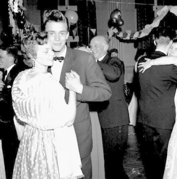 Ein Schwarz-Weiß-Foto eines Mannes und einer Frau, die auf einer Party tanzen, umgeben von lachenden Menschen und Ballons und Dekorationen im Hintergrund.