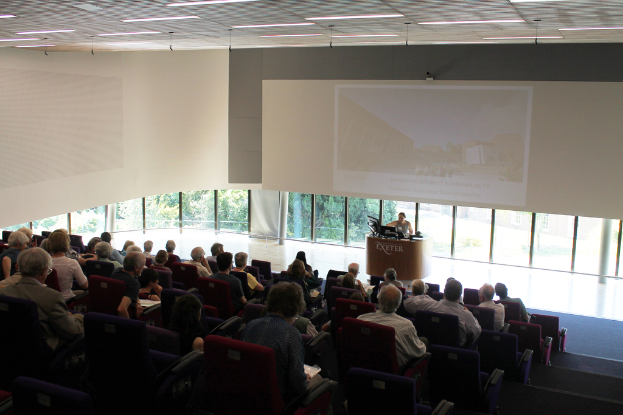 Ein Seminarsaal an einer Universität mit vielen Menschen, die aufmerksam einer Präsentation an der Front zugewandt sind, ein Redner steht an einem Tisch und erklärt die Präsentation, mit Glasfenstern im Hintergrund, die Bäume und Gebäude zeigen.