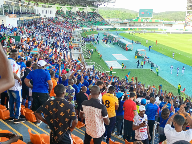 Eine große Menschenmenge versammelt sich vor einem eingezäunten Fußballfeld, wobei einige auf Stühlen sitzen und andere stehen, mit Blick auf Hügel unter einem klaren blauen Himmel während der African Cup of Nations 2019.