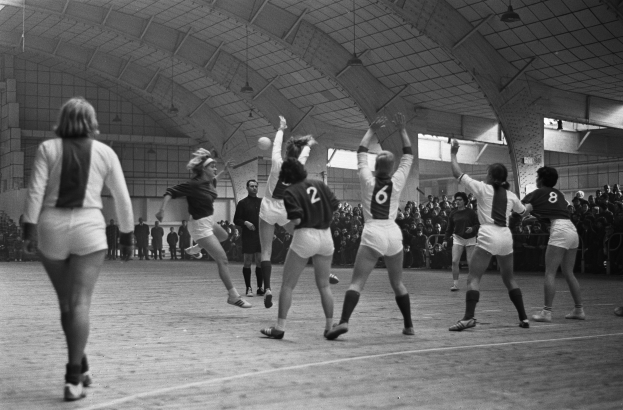 Eine Gruppe von Frauen beim Volleyballspielen in einer Turnhalle, mit Zuschauern auf Stühlen und Stehplätzen im Hintergrund, unter Deckenbeleuchtung.
