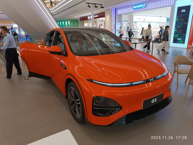 Orangefarbener Elektroauto in einer Ausstellungshalle umgeben von Menschen, Stühlen, einem Wagen und Läden im Hintergrund.