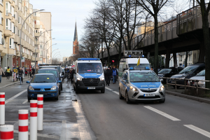 Eine Gruppe von Polizeiwagen fährt eine Straße mit hohen Gebäuden entlang, mit Menschen, die auf dem Bürgersteig gehen und Fahrräder fahren, eine Brücke mit Geländern auf der rechten Seite und Bäume mit einem klaren blauen Himmel im Hintergrund.