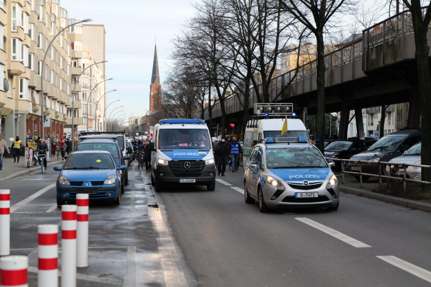 Eine Gruppe von Polizeiwagen fährt eine Straße mit hohen Gebäuden entlang, mit Menschen, die auf dem Bürgersteig gehen und Fahrräder fahren, eine Brücke mit Geländern auf der rechten Seite und Bäume mit einem klaren blauen Himmel im Hintergrund.