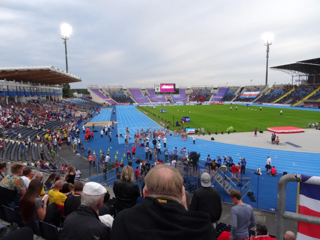 Großes Stadion voller Zuschauer bei einem Fußballspiel mit sitzenden und stehenden Fans, Geländern, einer Fahne, Flutlicht, einem Bildschirm, Bäumen und einem bewölkten Himmel.