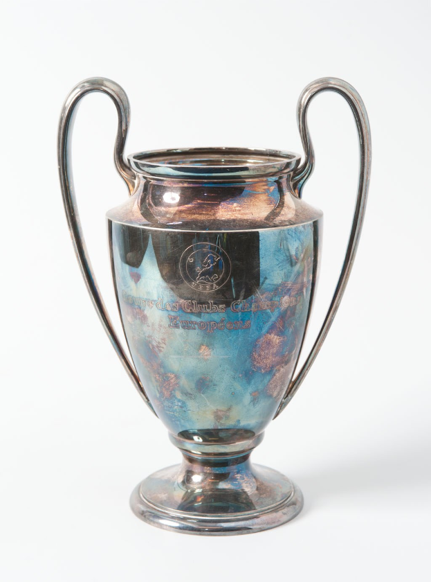 Der UEFA-Champions-League-Pokal, ein stählerner Becher mit zwei Griffen und einem blauen und silbernen Farbschema, der mit "UEFA Champions League" beschriftet ist und auf einem weißen Hintergrund steht.