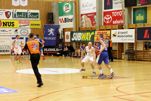 Ein Basketballfeld mit Spielern im Vordergrund und einer Holzwand mit Plakaten und Bannern im Hintergrund.