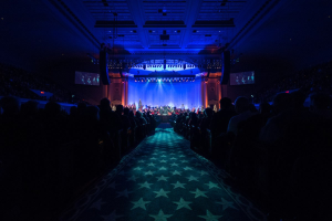 Ein großer Auditorium mit Sitzenden, eine Bühne mit Musikern und eine Decke mit Lichtern und Fernsehgeräten, was auf einen Konzertsaal für die Nationale Convention 2019 hindeutet.