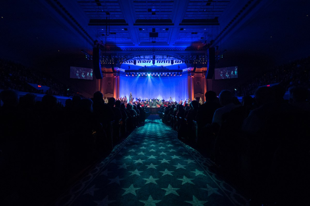 Ein großer Auditorium mit Sitzenden, eine Bühne mit Musikern und eine Decke mit Lichtern und Fernsehgeräten, was auf einen Konzertsaal für die Nationale Convention 2019 hindeutet.