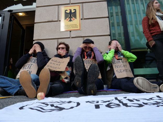 Eine Gruppe von Menschen sitzt vor einem Gebäude auf dem Boden und hält Schilder, mit einer Fahne auf der Straße und einer Frau rechts daneben, offenbar bei einer Protestaktion gegen eine Regierungsentscheidung.