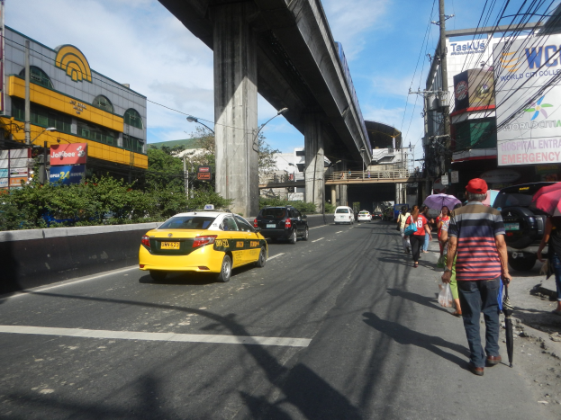 Ein gelbes Taxi fährt auf einer Stadtstraße mit hohen Gebäuden, Fußgängern mit Regenschirmen auf dem Gehweg, einer Brücke im Hintergrund, bewölktem Himmel und Strommasten mit Drähten.