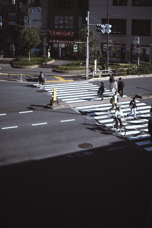 Eine Gruppe von Menschen überquert eine Straße an einer Ampel mit einem Radfahrer im Vordergrund und Gebäuden, Bäumen und Verkehrszeichen im Hintergrund.