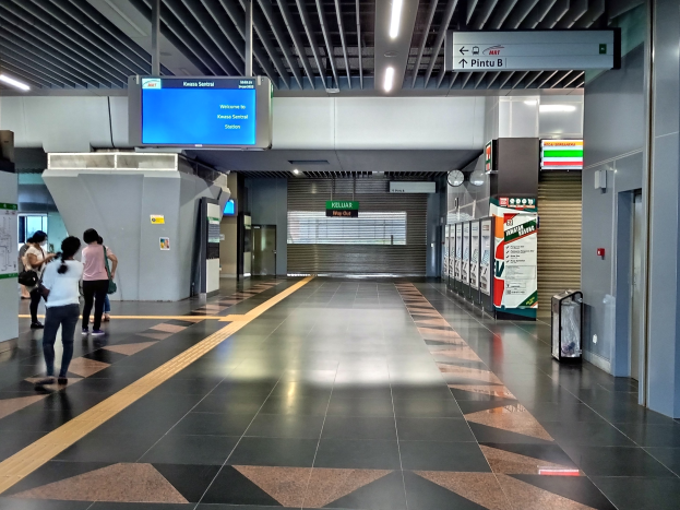 Menschen gehen durch einen Bahnhof mit einem Kofferwagen auf der rechten Seite, Schilder mit Text, eine Uhr an der Wand und Deckenleuchten.