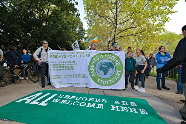Eine Gruppe von Menschen steht draußen und hält ein Banner mit der Aufschrift "Alle Flüchtlinge sind hier willkommen" und eine Flagge, mit Fahrrädern, einem Zaun, einem Straßenschild, einer Tafel, Bäumen und einem bewölkten Himmel im Hintergrund.