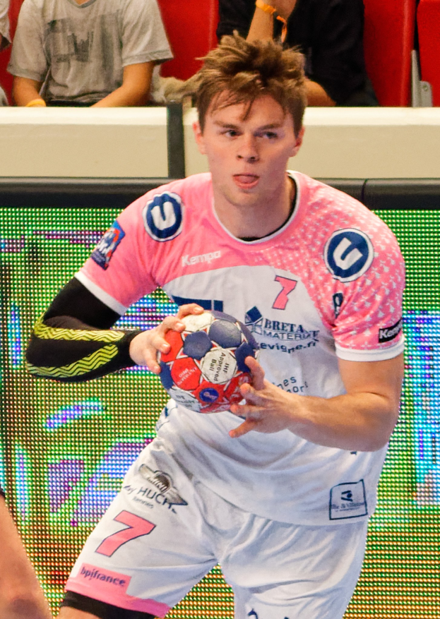 Ein Mann in einem pinken Hemd mit einem Fußball in der Hand, daneben ist die Hand einer anderen Person zu sehen, im Hintergrund sitzen mehrere Personen auf Stühlen.