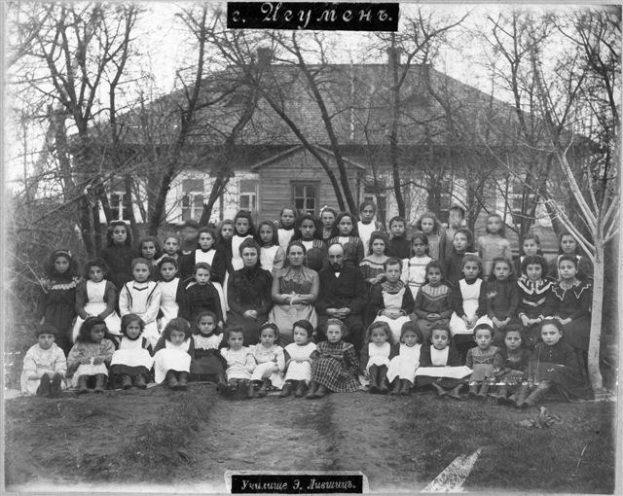 Schwarzes und weißes Foto einer Gruppe von Kindern, die vor einem Gebäude mit Fenstern posieren, umgeben von Bäumen und einem klaren Himmel, mit Text unten, der lautet: "Jugoslawien, die erste Schule der Welt."