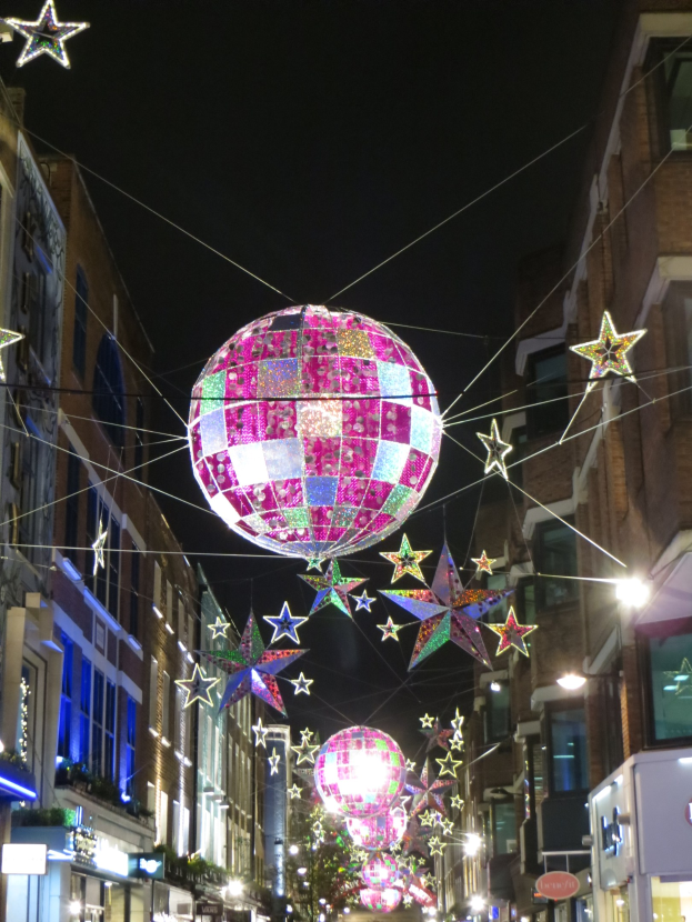 Eine Stadtstra├če bei Nacht mit festlicher Beleuchtung und Deko, darunter eine gro├če Diskokugel, die von einem Geb├Ąude h├Ąngt.