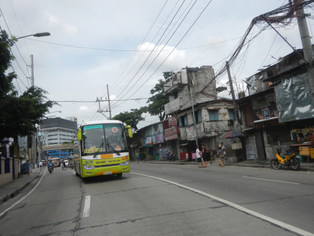 Ein grüner Bus fährt auf einer Straße mit Hochhäusern, Strommasten, Bäumen, Fußgängern und Radfahrern unter einem bewölkten Himmel.