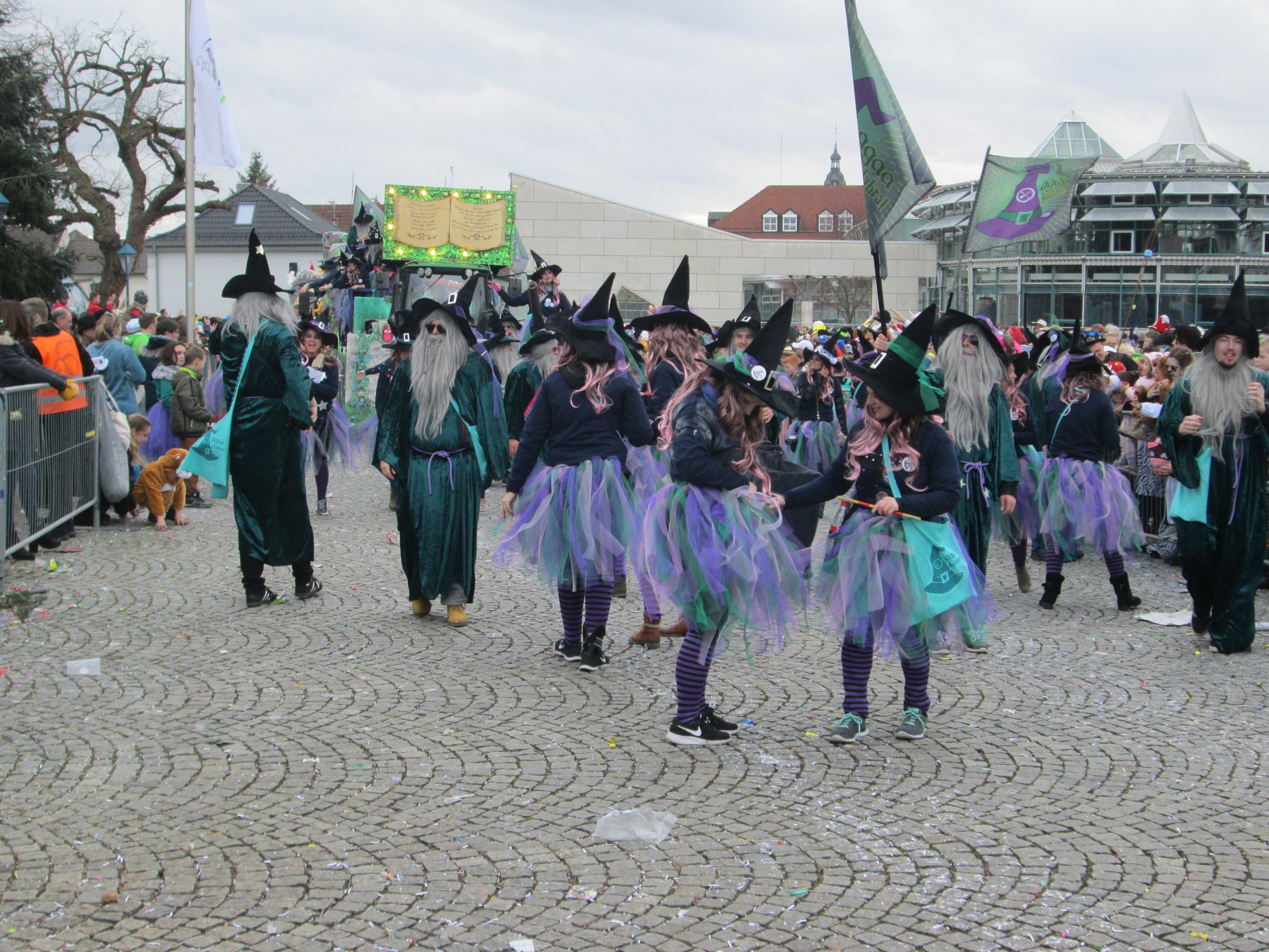 Gruppe von Menschen in Hexenkostümen, die über ein Kopfsteinpflasterstraße gehen und schwarze und lila Kleider und Hüte tragen, einige halten Fahnen, mit einem Geländer auf der linken Seite und Gebäuden, Bäumen und einem klaren blauen Himmel im Hintergrund.