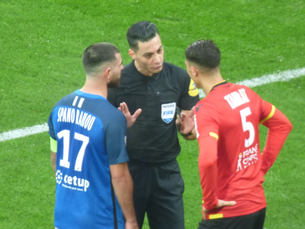 Schiedsrichter spricht mit zwei Männern auf einem Fußballfeld, einer trägt eine schwarze Hose und ein schwarzes Hemd, der andere ein blaues und rotes T-Shirt mit Text und Brille.