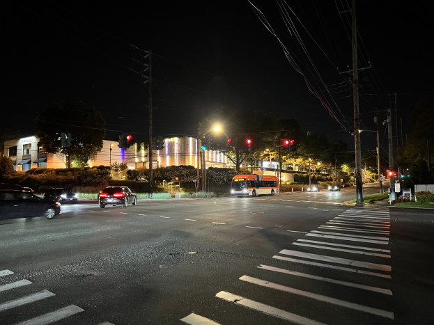 Eine nächtliche Stadtstraße mit fahrenden Autos und einem Bus, beleuchtet von Straßenlaternen und Ampeln, umgeben von Gebäuden, Bäumen und Gras unter einem dunklen Himmel.