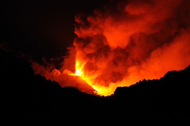 Ein Vulkan bricht nachts aus, mit leuchtend orangefarbenem und gelbem Lavafluss ins Meer und Rauch, der in den dunklen Himmel aufsteigt.