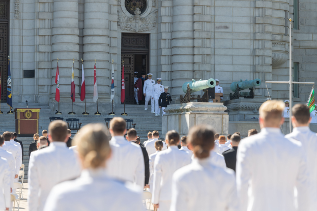 Gruppe von Menschen in weißen Marineuniformen, die auf einer Treppe vor einem Gebäude mit Säulen, einem Podium mit Mikrofon und Fahnen während einer Abschlusszeremonie stehen.