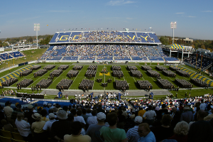 Eine große Menschenmenge in einem Stadion bei einem Fußballspiel, wobei einige stehen und andere sitzen, auf einem Rasenfeld unter einem klaren blauen Himmel, umgeben von Laternenmasten, Bäumen und Gebäuden.