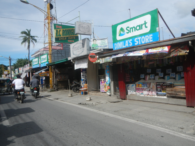 Eine belebte Straße mit Motorradfahrern, Autos, Gebäuden, Schildern, Strommästen, Kabeln, Straßenlaternen, Bäumen und einem bewölkten Himmel.