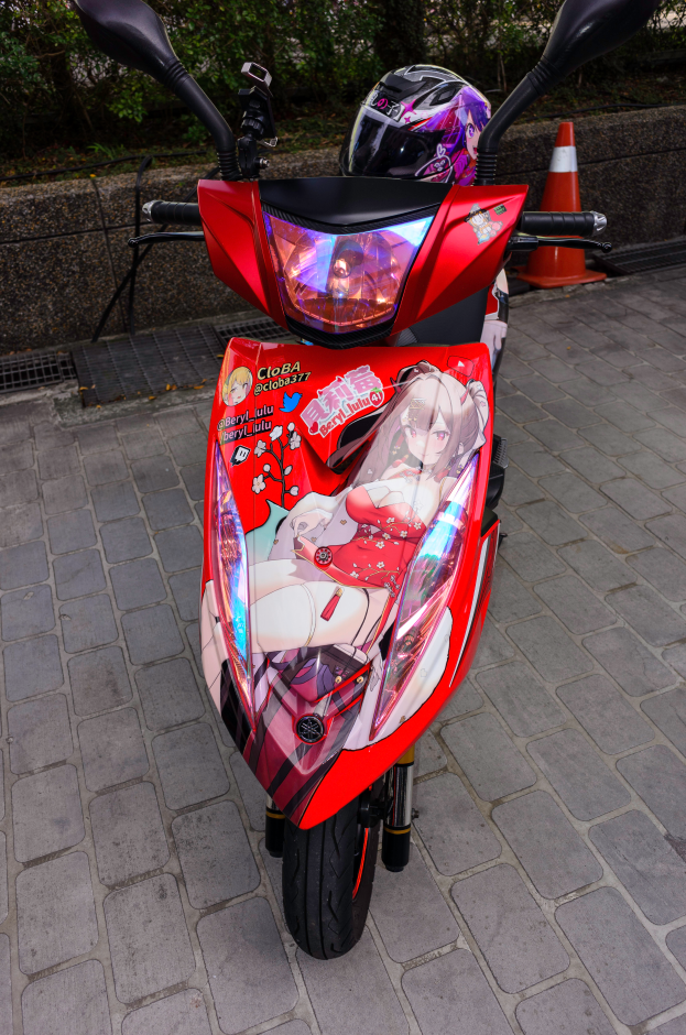Ein oranger Roller am Straßenrand mit einem Helm daneben, mit einer Verkehrskegel, einer Wand und mehreren Bäumen im Hintergrund.