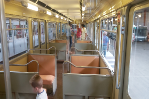 Eine Gruppe von Menschen, die auf einem Zug mit Sitzen, Pfosten und Glasfenstern fahren, mit einem Jungen im Vordergrund und anderen Fahrzeugen außerhalb sichtbar.