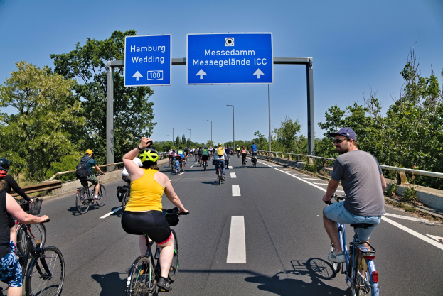 Eine Gruppe von Radfahrern mit Helmen fährt auf einer Straße mit einer Begrenzung auf einer Seite und Bäumen auf der anderen, unter einem klaren blauen Himmel mit Laternen im Hintergrund.