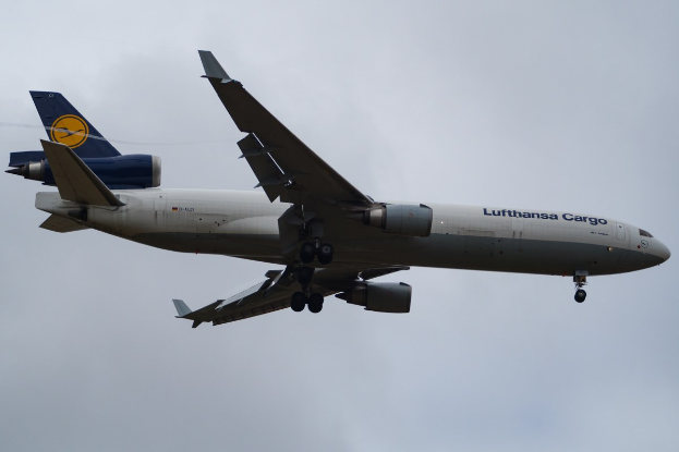 Ein Lufthansa Cargo Boeing 777-300ER im Flug, mit ausgebreiteten Tragflächen und aktivierten Triebwerken, mit sichtbarem Text auf dem Rumpf gegen einen teilweise bewölktem Himmel.