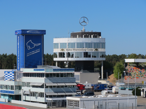 Eine Ansicht des Mercedes-AMG Petronas F1 Team-Hauptquartiers in Berlin, Deutschland, mit Gebäuden, Fahrzeugen auf der Straße, einem Zaun, Pfählen, Schildern mit Text, Bäumen und einem Logo auf dem Gebäude, mit dem Himmel im Hintergrund.