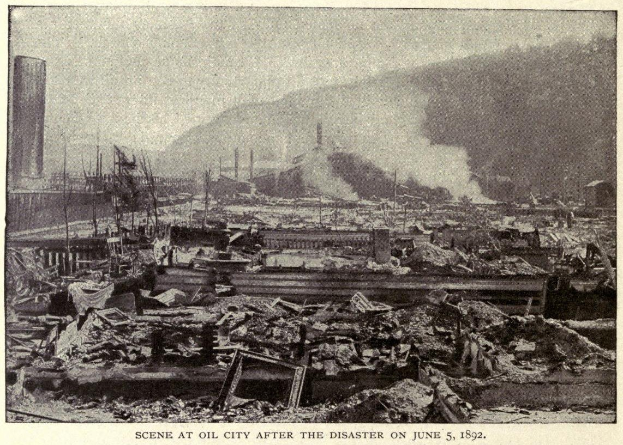 Schwarzes und weißes Foto einer Stadt nach einem Unglück am 5. Juni 1892, das Gebäude, Pfähle, Rauch und einen Hügel im Hintergrund zeigt, mit Text unten.