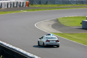 Polizeiauto auf einer Rennstrecke mit Gras auf beiden Seiten, Menschen und Zelte im Hintergrund sichtbar.