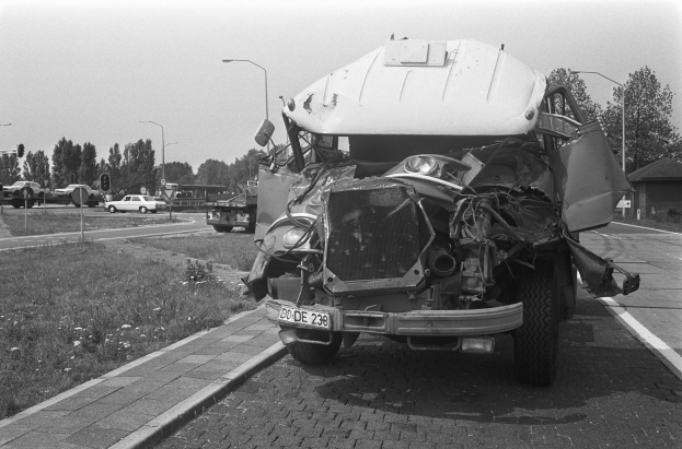 Ein Schwarz-Weiß-Foto eines stark beschädigten Autos am Straßenrand mit eingedellter Motorhaube und zersplitterten Fenstern und anderen Fahrzeugen, Laternenpfählen, Bäumen und einem Haus im Hintergrund.
