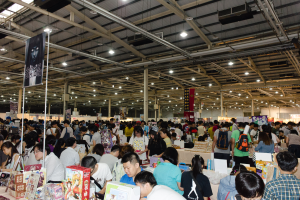 Eine große Gruppe von Menschen versammelt sich um Tische, die mit Büchern, Kartons und anderen Gegenständen gefüllt sind, auf einer Buchmesse mit Bannern und Lichtern oben.