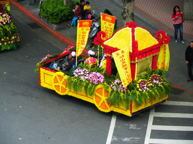Schwimmend in einem mit Blumen und Bannern geschmückten Paradewagen, der Reiter auf dem Dach trägt, mit Zuschauern auf dem Gehweg rechts und Bäumen im Hintergrund.