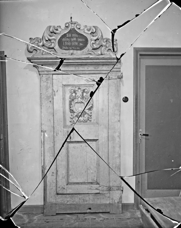 Ein Schwarz-Weiß-Foto einer zerbrochenen Tür in einem Raum mit einer Wand im Hintergrund, die Tür in einem schlechten Zustand mit verstreuten Glasscherben.