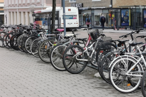 Eine Reihe von Fahrrädern, die an der Straße geparkt sind, mit einem Bus im Hintergrund, Menschen auf dem Gehweg, ein Baum und Gebäude mit Fenstern in der Ferne.