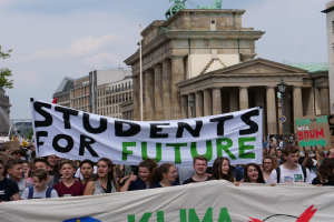 Eine Gruppe von Schülern marschiert in Berlin, hält ein buntes "Students for Future"-Schild gegen den Hintergrund von Gebäuden, Bäumen und Himmel.