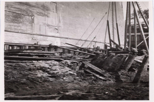 Ein Schwarz-Weiß-Foto einer Baustelle für das New Yorker Staatsgefängnis, das Arbeiter, hölzerne Gegenstände, Pfähle, Seile, eine Wand und Bäume im Hintergrund zeigt.