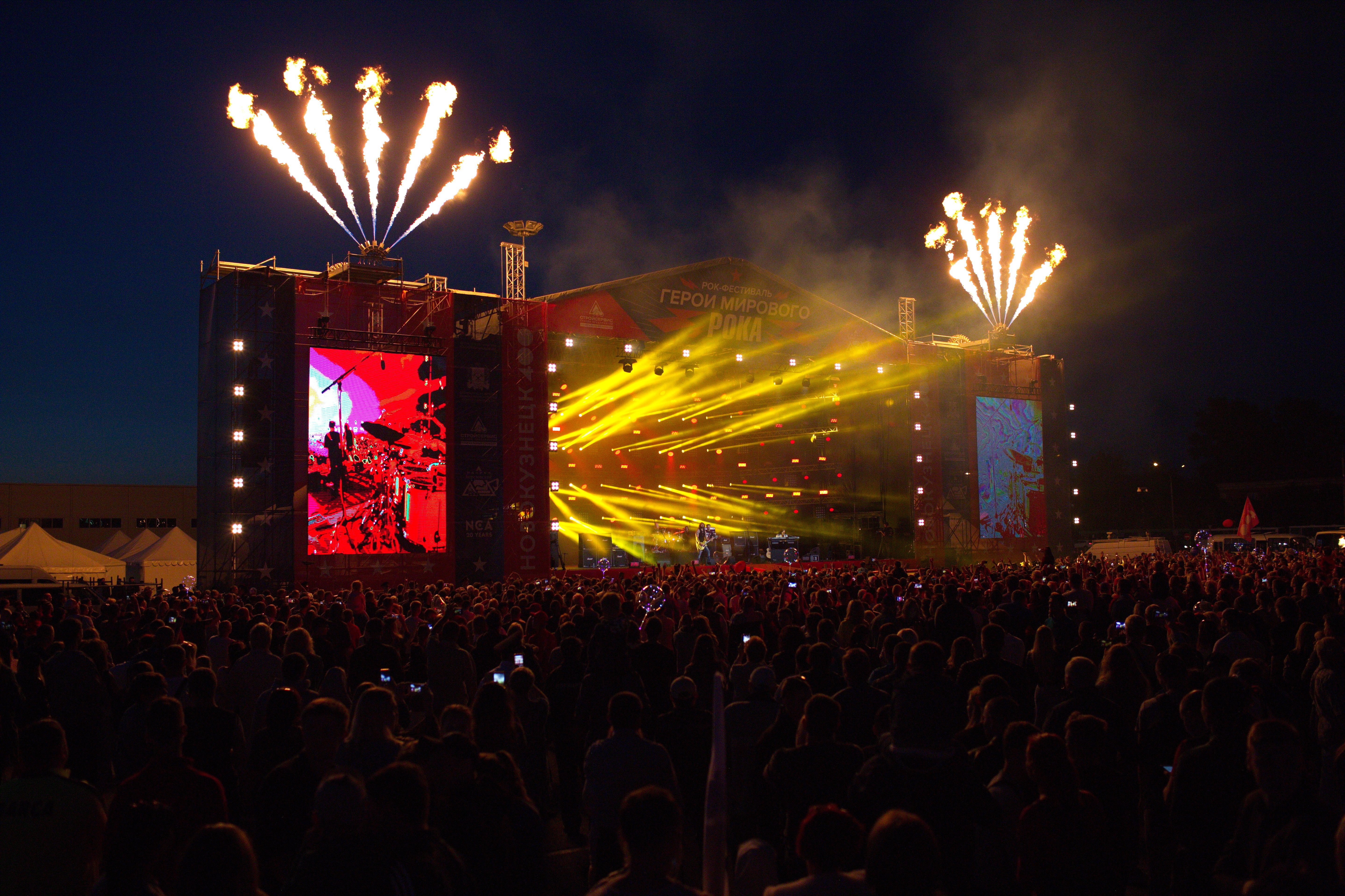 Eine große Menschenmenge versammelte sich auf einem Musikfestival, mit einer Bühne in der Mitte, auf der Musiker Instrumente spielen, beleuchtet von hellen Lichtern und umgeben von Bannern, Zelten links und einem dunklen Himmel mit Feuerwerk im Hintergrund.