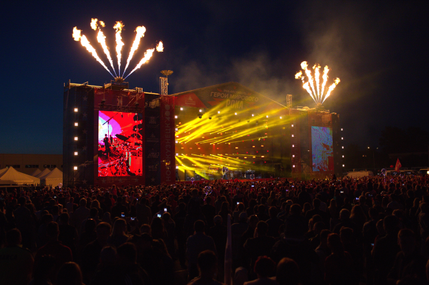 Eine große Menschenmenge versammelte sich auf einem Musikfestival, mit einer Bühne in der Mitte, auf der Musiker Instrumente spielen, beleuchtet von hellen Lichtern und umgeben von Bannern, Zelten links und einem dunklen Himmel mit Feuerwerk im Hintergrund.