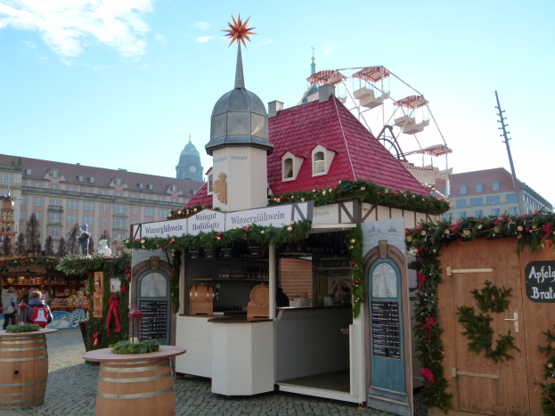 Kleines rotes Dachgebäude mit einem Riesenrad davor, umgeben von festlichen Oktoberfest-Dekorationen, Menschen und Gegenständen, vor einem Hintergrund aus Gebäuden, Bäumen und einem bewölkten Himmel in München, Deutschland.
