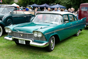 Alte Autos auf Rasen mit Menschen davor, Zelte im Hintergrund, Bäume oben und eine Flagge oben links.