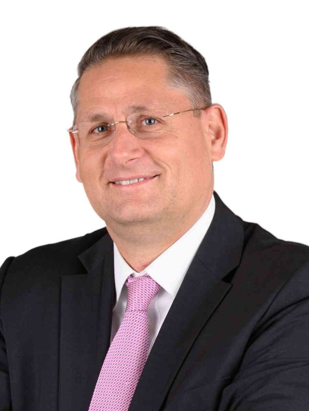 Ein lachender Mann im Anzug und Krawatte, mit Brille, vor einem weißen Hintergrund.