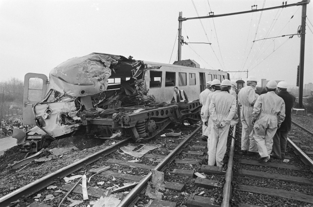 Schwarzes und weißes Bild einer Zugkollision auf den Schienen mit Menschen in Helmen in der Nähe, umgeben von Strommasten, Bäumen, Gebäuden und einem klaren Himmel.