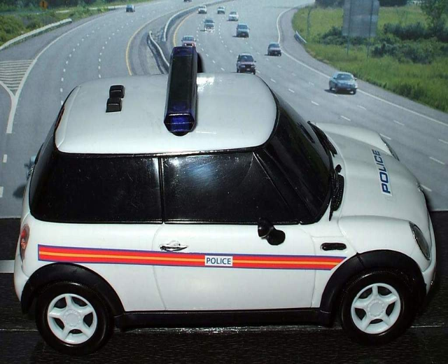 Weißes Polizeiauto mit "Polizei" Aufschrift vor bewegendem Verkehr auf einer Straße gesäumt von Gras und Bäumen außerhalb einer Stadt.