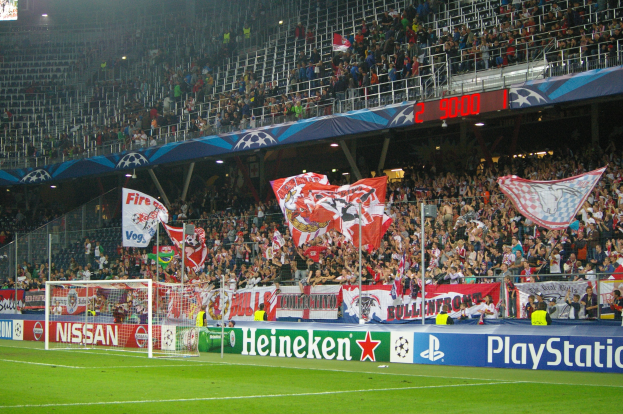 Ein Fußballspiel in einem hell erleuchteten Stadion mit einer großen Zuschauermenge, grünem Rasen, einem Tor, Bannern und einem Bildschirm in der linken oberen Ecke.
